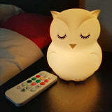 Veilleuse hibou à LED allumée en blanc, placée près d’un lit, avec sa télécommande posée à côté
